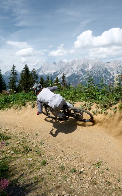 Ein Mountainbiker driftet staubend durch eine Kurve der Rookie Downhill Strecke mit Blick zum Dachstein. | © Dietmar Koerbler