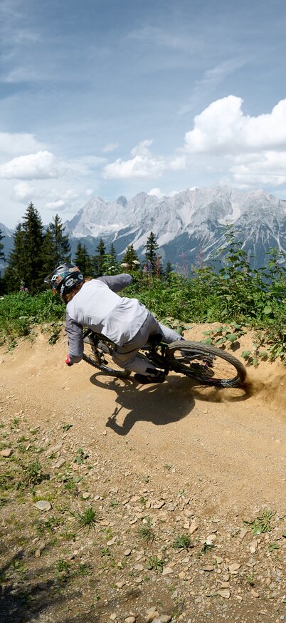 Ein Mountainbiker driftet staubend durch eine Kurve der Rookie Downhill Strecke mit Blick zum Dachstein. | © Dietmar Koerbler