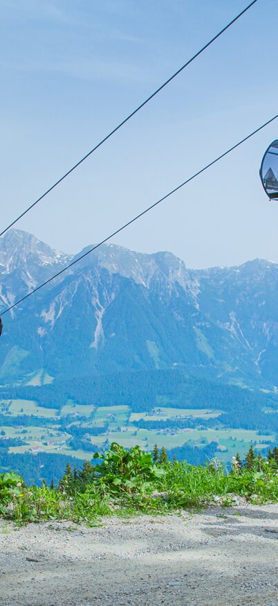 Familie bestehend aus Vater, kleinem Kind in der Mitte und Mutter gehen entlang eines Weges bei blauem Himmel mit Panorama und einer Planai Gondel im Hintergrund. | © René Eduard Perhab