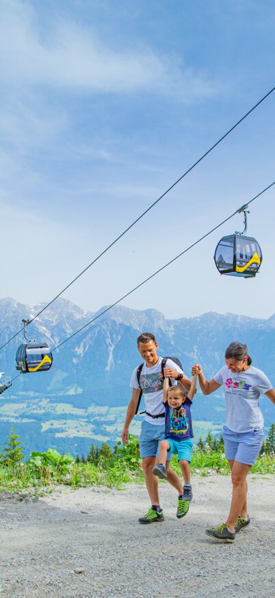 Familie bestehend aus Vater, kleinem Kind in der Mitte und Mutter gehen entlang eines Weges bei blauem Himmel mit Panorama und einer Planai Gondel im Hintergrund. | © René Eduard Perhab