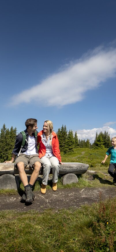 Familie mit zwei Kindern wandert auf den Berg und macht Rast auf einer Bank mit Ausblick auf die Berge | © Tom Lamm | ikarus.cc