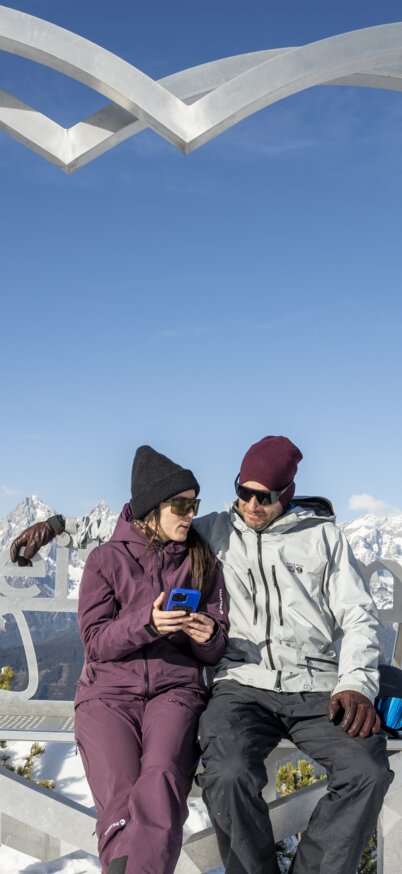 Paar mit Handy in der Hand sitzt auf dem Steirerherz, ein großes Herz aus Stahl, auf der Reiteralm. | © Lorenz Masser