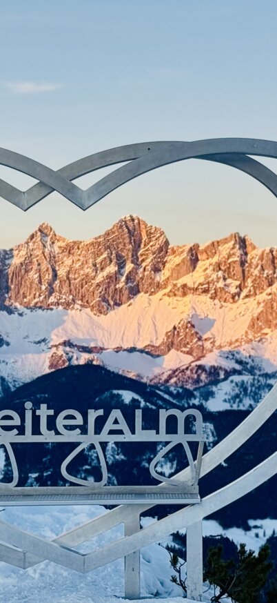 Steirerherz mit Schriftzug Reiteralm in der Mitte steht am Gipfel der Gasselhöhe auf der Reiteralm mit Ausblick auf das verschneite Dachsteinmassiv. | © Reiteralm Bergbahnen