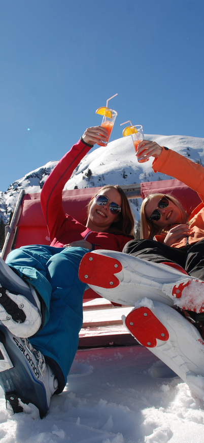 2 junge Skifahrerinnen sitzen auf 2 Liegestühlen im Schnee auf der Reiteralm und stossen mit ihren Getränken zusammen. | © Reiteralm Bergbahnen