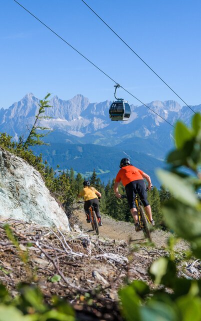 2 Biker sind auf den Reiteralm Trails unterwegs. Im Hintergrund das Dachsteinmassiv und eine Gondelkabine des Preunegg Jets auf der Reiteralm. | © Matthias Fritzenwallner
