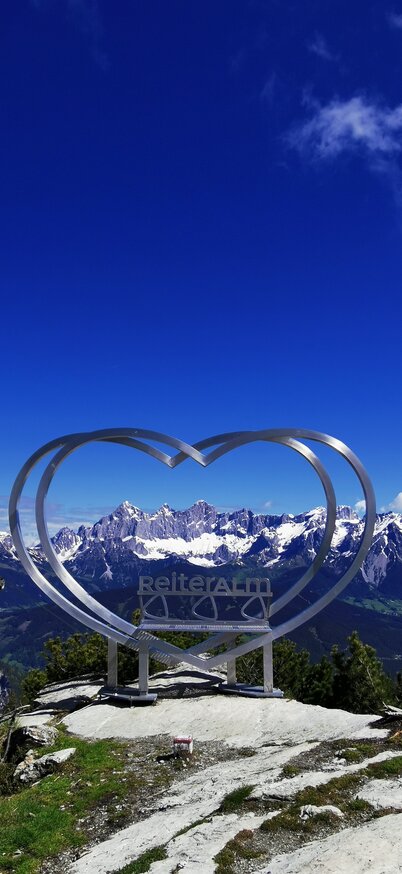 Steirerherz mit Schriftzug Reiteralm in der Mitte steht am Gipfel der Gasselhöhe auf der Reiteralm mit Ausblick auf das verschneite Dachsteinmassiv. | © Reiteralm Bergbahnen