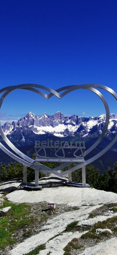 Steirerherz mit Schriftzug Reiteralm in der Mitte steht am Gipfel der Gasselhöhe auf der Reiteralm mit Ausblick auf das verschneite Dachsteinmassiv. | © Reiteralm Bergbahnen