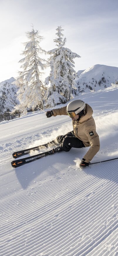 Ein Skifahrer fährt auf der Piste bergab und zieht dabei eine Schneewolke mit sich, im Hintergrund sieht man eine weite Gipfellandschaft im Schnee und Bäume | © Mirja Geh