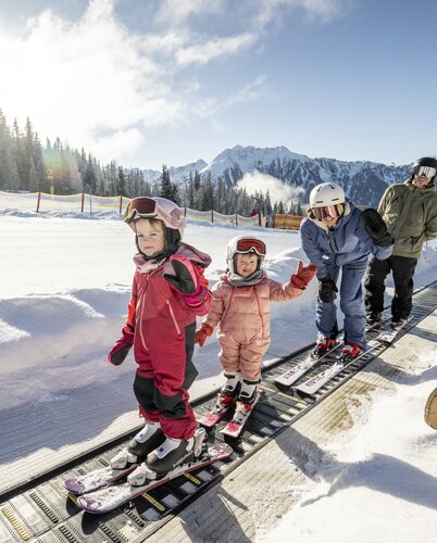 Zwei Kinder und zwei Erwachsene, gekleidet mit einem Skianzug, benutzen ein Förderband, beide Kinder und eine Erwachsene winken in die Kamera, der zweite Erwachsene schaut in die Kamera, im Hintergrund sieht man einen Zaun, Schnee sowie Wald, blauen Himmel und eine große Wolke | © Lorenz Masser