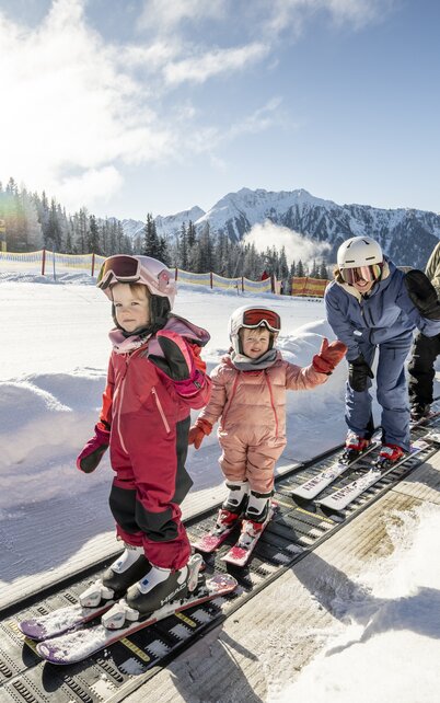 Zwei Kinder und zwei Erwachsene, gekleidet mit einem Skianzug, benutzen ein Förderband, beide Kinder und eine Erwachsene winken in die Kamera, der zweite Erwachsene schaut in die Kamera, im Hintergrund sieht man einen Zaun, Schnee sowie Wald, blauen Himmel und eine große Wolke | © Lorenz Masser