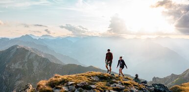 Zwei Personen die auf einem Berggrat entlang wandern, im Hintergrund sieht man weite Berglandschaft, ein paar Wolken und die durchscheinende Sonne | © Mathäus Gartner