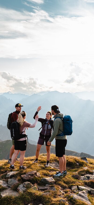 Vier Personen stehen auf einem Gipfel und zwei davon klatschen miteinander ein, im Hintergrund sieht man weite Berglandschaft | © Mathäus Gartner