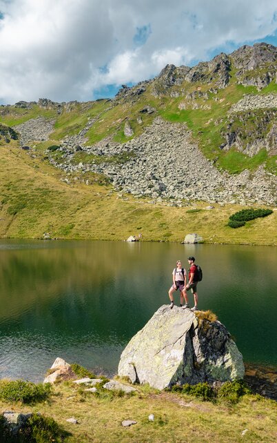 Zwei Personen stehen auf einem großen Stein, der sich direkt an einem Bergsee befindet, der Bergsee wird von Bergen umgeben  | © Mathäus Gartner