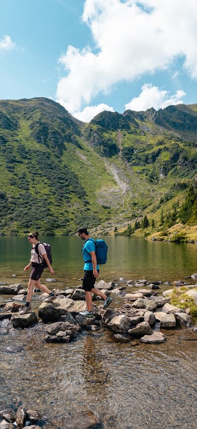 Zwei Personen balancieren auf Steinen durch einen Bergsee, der von hohen Bergen umgeben ist | © Mathäus Gartner