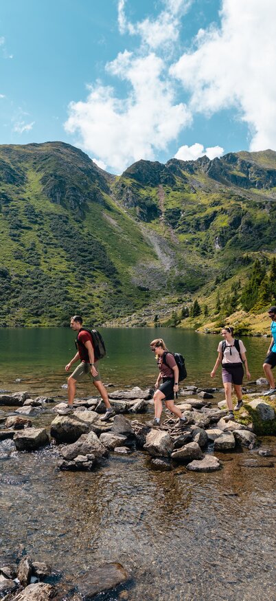 Vier Personen balancieren auf Steinen durch einen Bergsee, der von hohen Bergen umgeben ist | © Mathäus Gartner