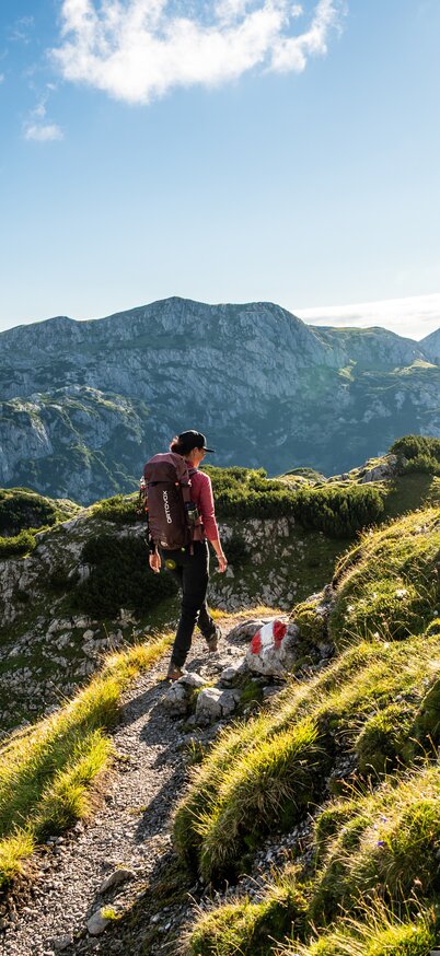 Eine Dame wandert einen schmalen Weg entlang, im Hintergrund sieht man Berge, Wolken und blauen Himmel | © Janna Kamphof