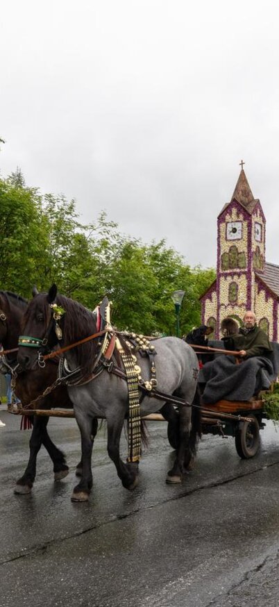 Ein festlicher Umzug mit einem Pferdegespann, das einen kunstvoll dekorierten Wagen zieht. Im Hintergrund ist eine kleine Kirche und eine grüne Landschaft zu sehen. | © Michael Simonlehner