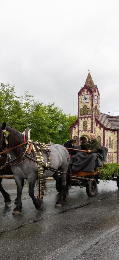 Ein festlicher Umzug mit einem Pferdegespann, das einen kunstvoll dekorierten Wagen zieht. Im Hintergrund ist eine kleine Kirche und eine grüne Landschaft zu sehen. | © Michael Simonlehner