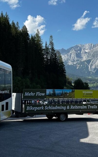 Ein Bus steht auf einem Parkplatz vor majestätischen Bergen und einer grünen Waldlandschaft. Im Hintergrund sind wolkiger Himmel und ein Eintrag für den Bikepark Schladming sichtbar. | © TVB Schladming-Dachstein